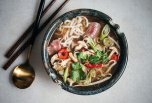A bowl of noodle soup with slices of raw beef, mushrooms, red chili, green onions, cilantro, and rich pho broth is served next to chopsticks and a gold spoon on a light surface.
