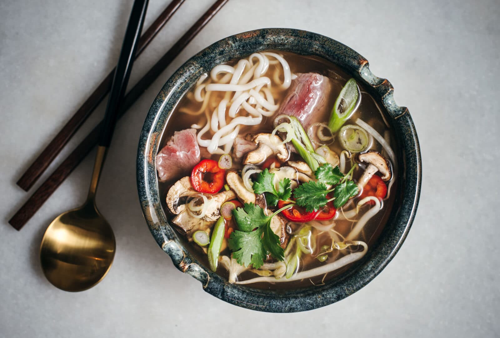 A bowl of noodle soup with slices of raw beef, mushrooms, red chili, green onions, cilantro, and rich pho broth is served next to chopsticks and a gold spoon on a light surface.