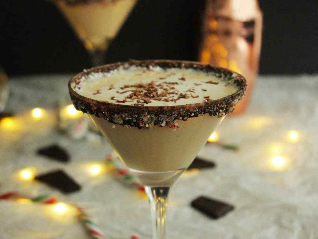 A creamy cocktail in a martini glass with a chocolate and sprinkle rim, topped with chocolate shavings. Festive lights, candy canes, and chocolate pieces are blurred in the background.