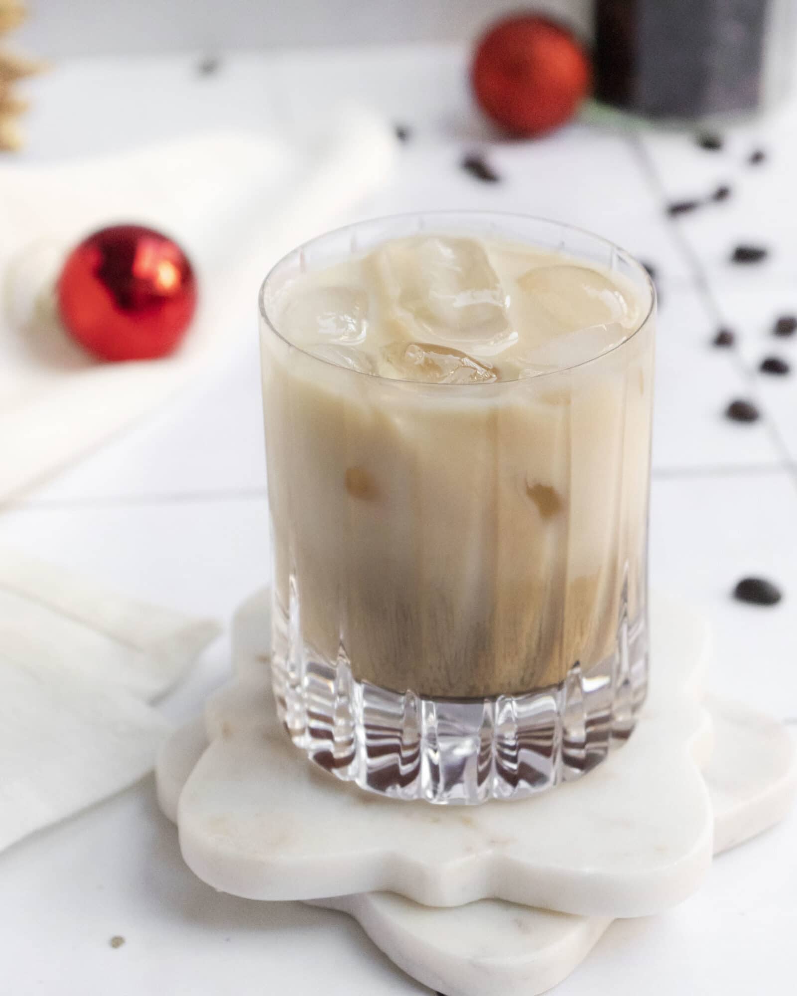 A glass of iced creamy coffee sits on a white marble coaster shaped like a flower, with red ornaments and chocolate chips scattered in the blurred background.
