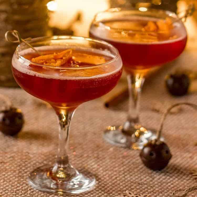 Two coupe glasses filled with a red cocktail, each garnished with orange slices on a pick, set on a textured surface with festive lights and decorative ornaments in the background.