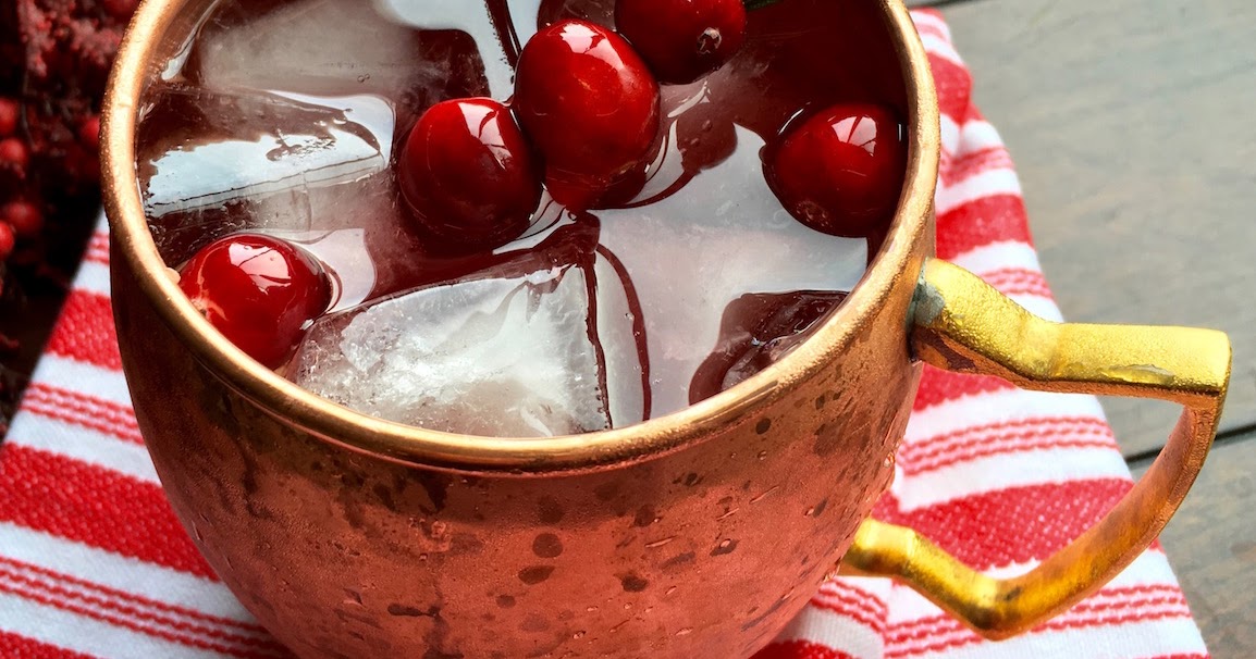 A copper mug filled with a red drink, ice cubes, and fresh cranberries, resting on a red and white striped cloth.