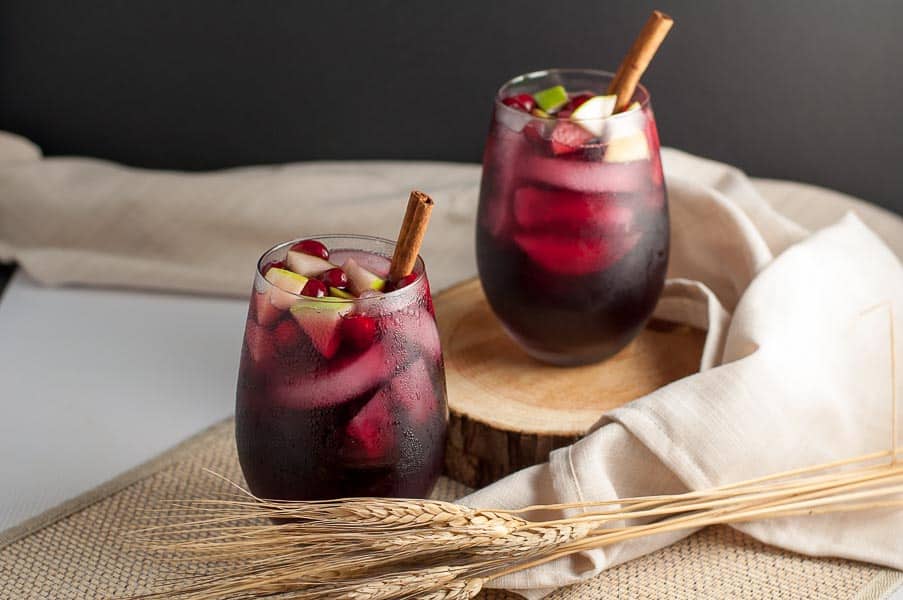 Two glasses of red sangria with ice cubes, diced apples, and cinnamon sticks, placed on a wooden surface with a beige cloth and wheat stalks for decoration.