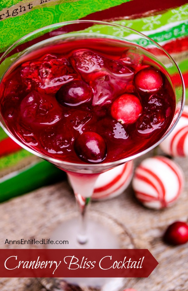 A martini glass filled with a red cranberry cocktail, ice, and whole cranberries, set against a festive background with red, white, and green holiday decorations. Text on the image reads “Cranberry Bliss Cocktail.”.