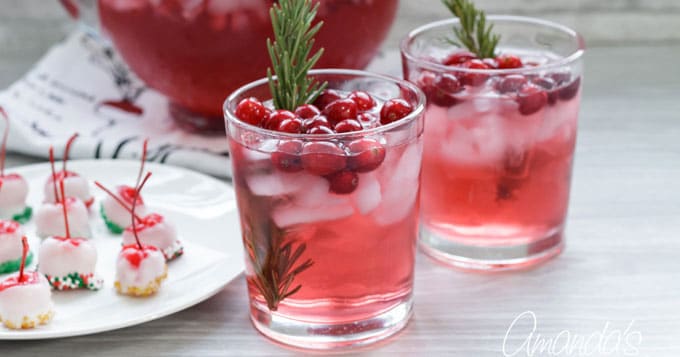 Two glasses of pink cranberry cocktail with ice, cranberries, and rosemary sprigs; a plate of cherry-topped treats and a punch bowl are in the background, all on a light-colored surface.