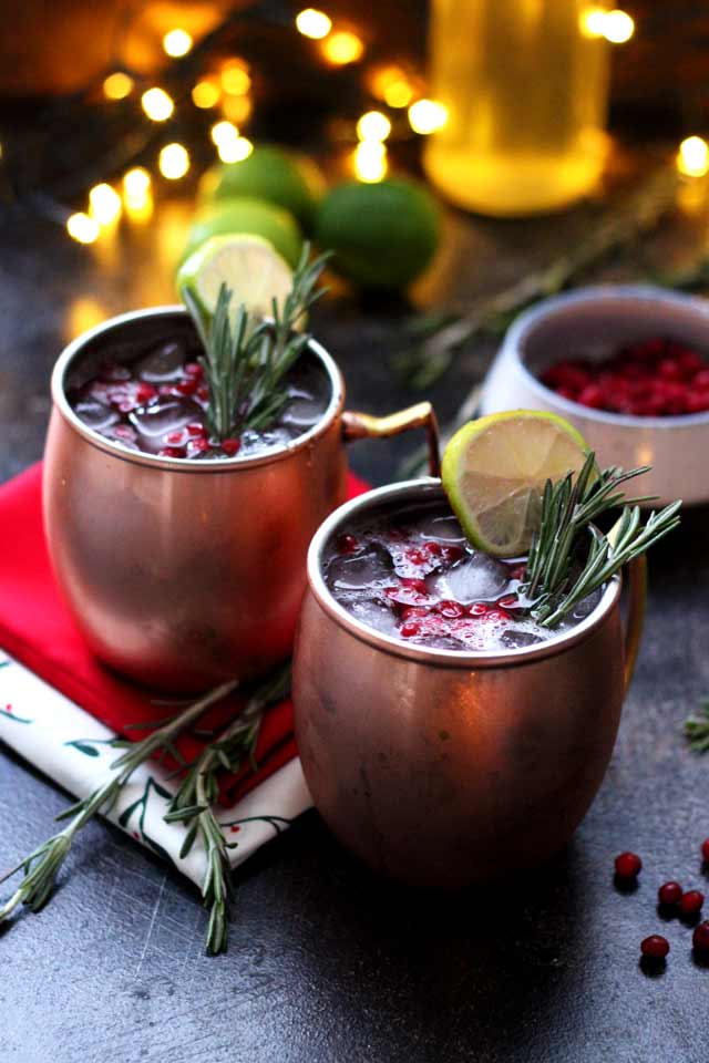 Two copper mugs filled with a festive drink, garnished with lime slices, rosemary sprigs, and red berries. A red napkin, limes, berries, and twinkling lights are in the background.