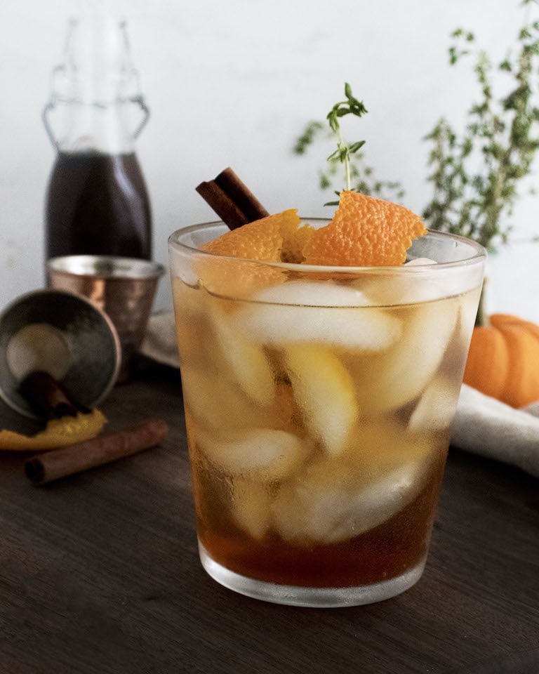 A glass filled with ice, amber liquid, orange peel, a cinnamon stick, and a herb garnish sits on a wooden table, with a bottle, a jigger, and cinnamon sticks in the background.