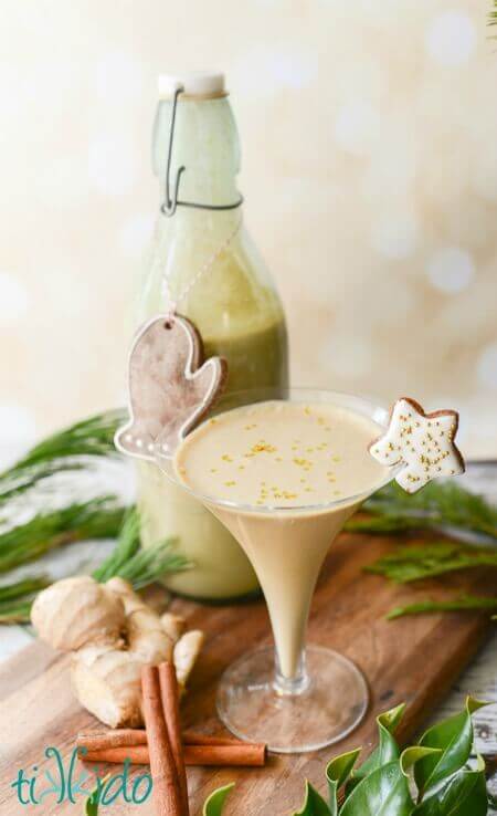 A martini glass filled with a creamy drink, topped with gold sprinkles and a star-shaped cookie on the rim, sits beside a bottle of similar drink, ginger root, and cinnamon sticks on a wooden board with greenery.