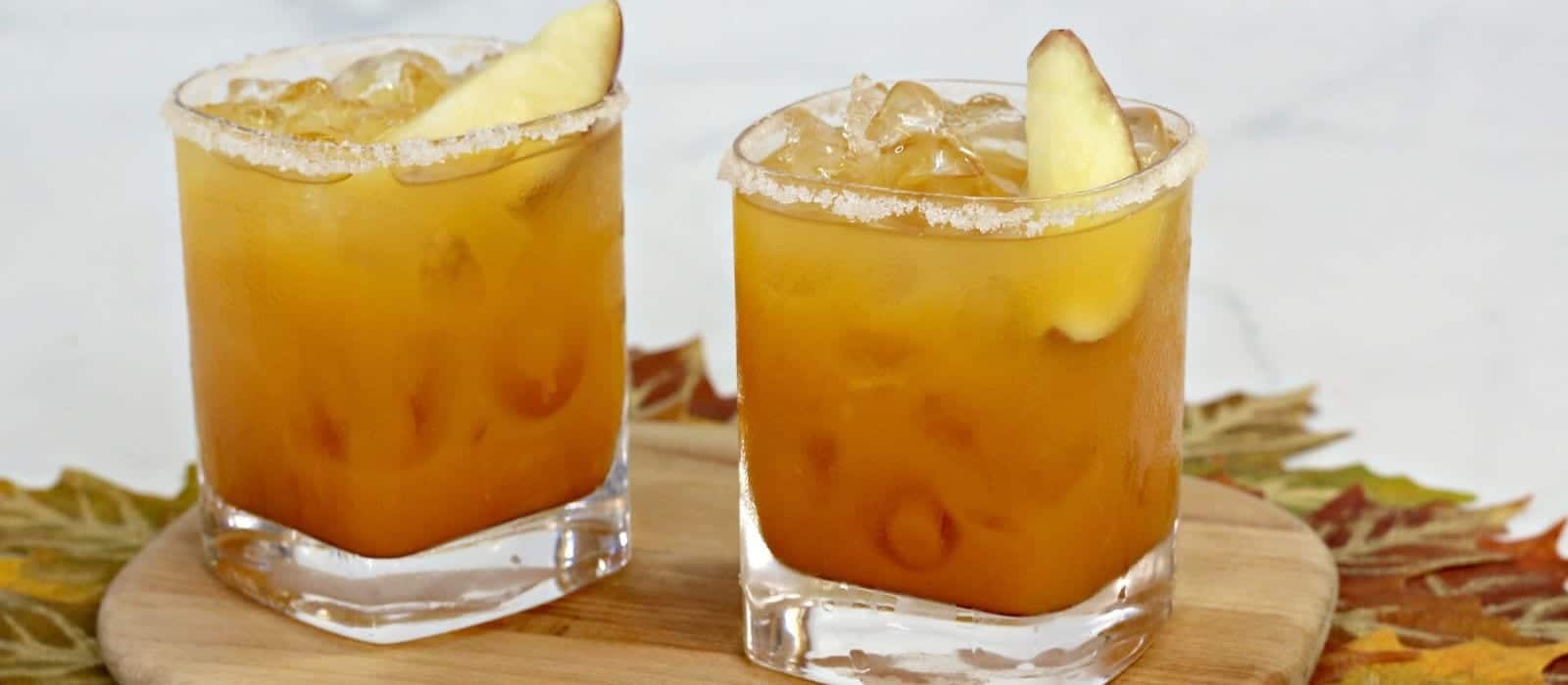 Two square glasses filled with orange-colored iced drinks, garnished with apple slices on the rim, sit on a wooden board with autumn leaves in the background.