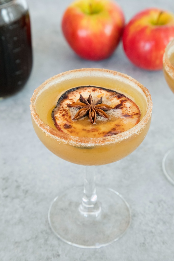 A cocktail in a glass with a cinnamon-sugar rim, garnished with a slice of bruleed apple and a star anise pod. Two red apples and a jar are blurred in the background.