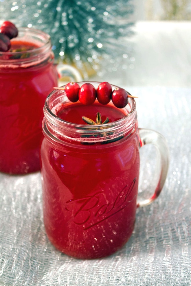 A mason jar filled with a bright red drink, garnished with whole cranberries on a skewer and star anise, sits on a textured silver surface with another similar jar and a festive background.