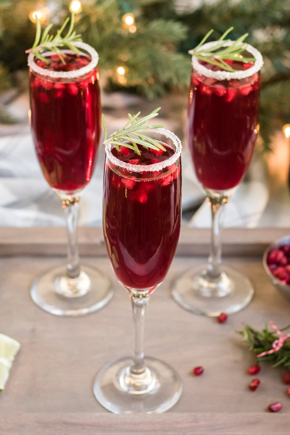 Three champagne flutes filled with a deep red beverage, garnished with sprigs of rosemary, sugared rims, and pomegranate seeds, set on a tray with festive greenery in the background.