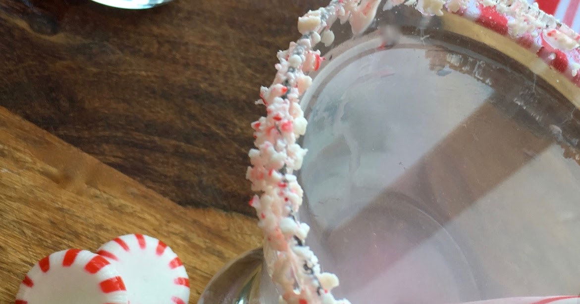 A glass with its rim coated in crushed peppermint candy, sitting on a wooden surface next to two whole peppermint candies.