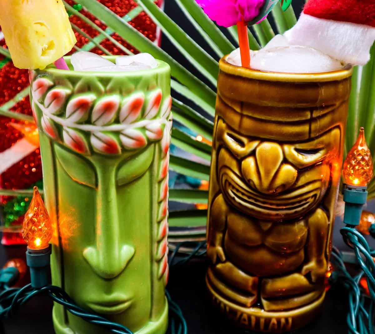 Two colorful tiki mugs with tropical cocktails, one green with a face design and one brown with a carved figure, are surrounded by palm leaves, orange string lights, and holiday decorations.