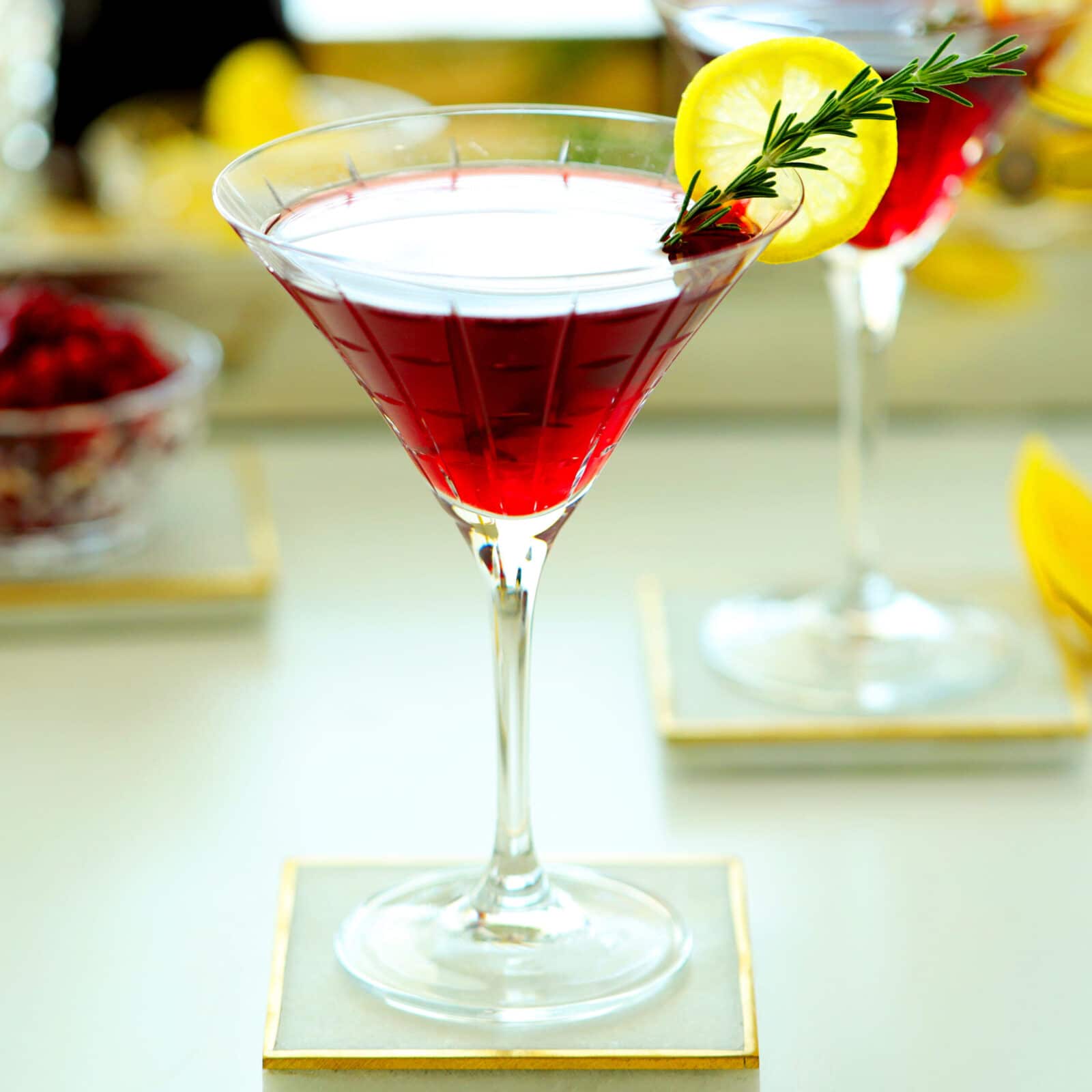 A red cocktail in a martini glass, garnished with a lemon slice and a sprig of rosemary, sits on a square coaster with another similar drink and blurred background elements.