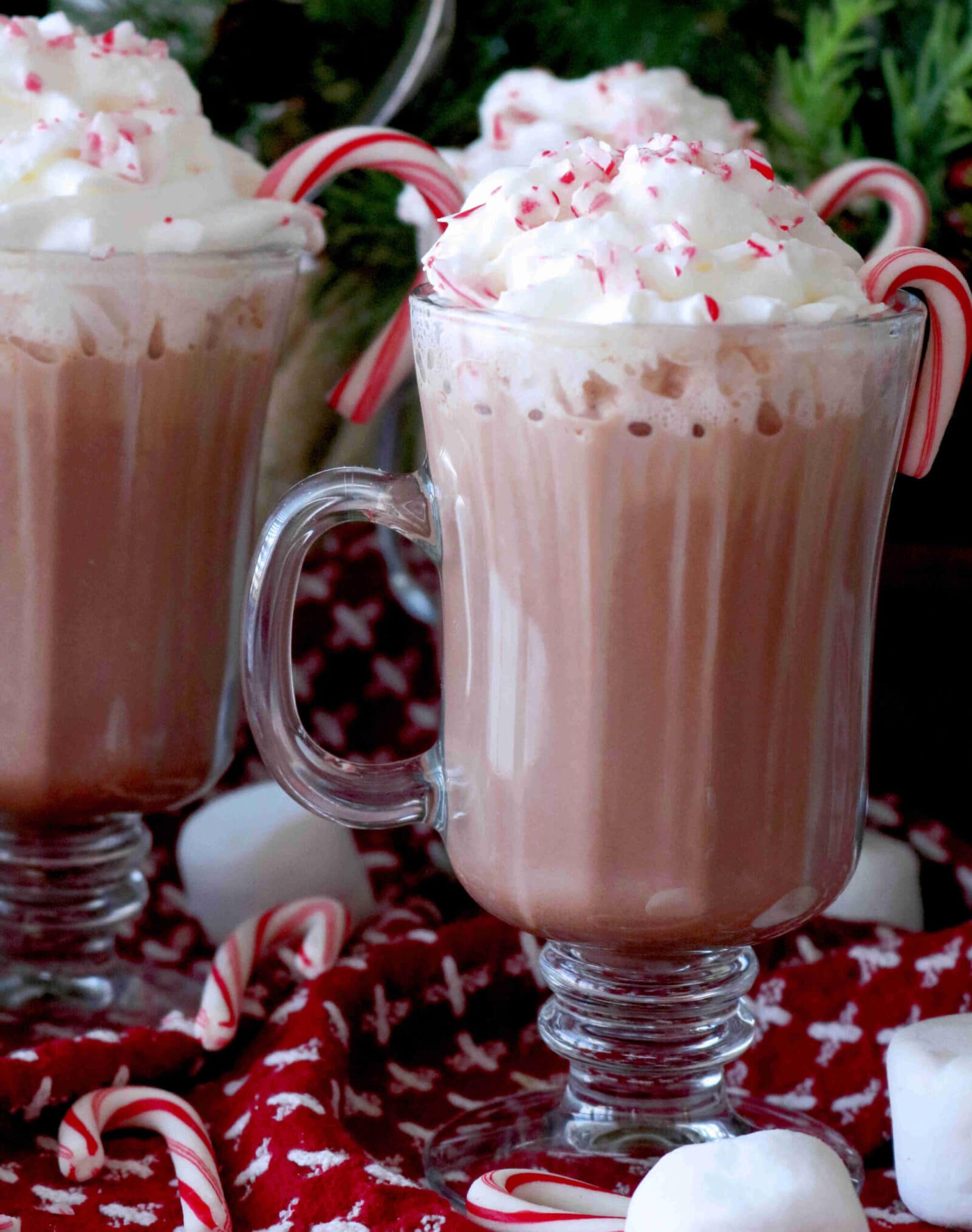 Two glass mugs of hot chocolate topped with whipped cream and crushed peppermint, each garnished with a candy cane. Marshmallows and mini candy canes are scattered on a red cloth with white patterns.