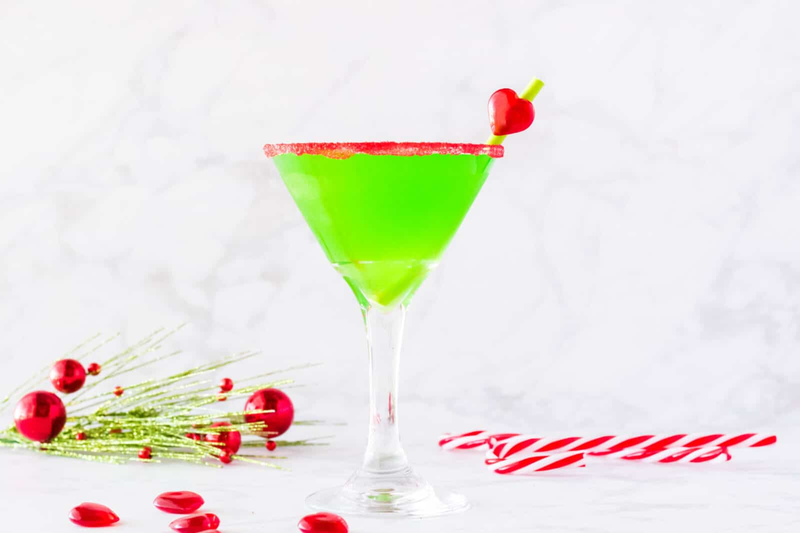 A bright green cocktail in a martini glass with a red sugar rim, garnished with a cherry and heart-shaped candy. Red ornaments, glittery greenery, and a striped candy cane are nearby on a white surface.
