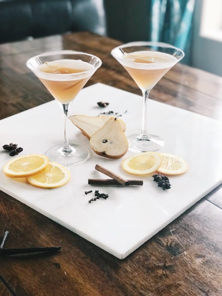 Two martini glasses filled with a light brown cocktail sit on a marble board, surrounded by pear slices, orange slices, cinnamon sticks, star anise, and cloves on a wooden table.