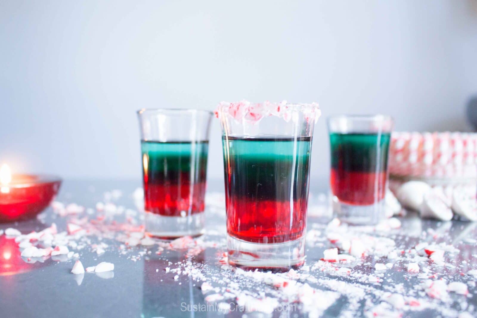 Three layered holiday shots in clear glasses, with red, green, and clear layers, stand on a surface scattered with crushed peppermint candy; one glass has a crushed peppermint rim. A lit candle and candy are in the background.