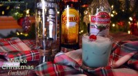A glass of White Christmas cocktail with ice and a cherry garnish sits on a red plaid cloth, surrounded by bottles of Smirnoff vodka, Kahlúa, and DeKuyper, in front of a lit Christmas tree. Text reads: “White Christmas Cocktail.”.