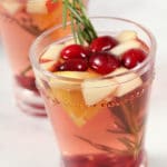 A glass of pink drink garnished with fresh cranberries, apple chunks, orange pieces, and a sprig of rosemary, with another similar glass blurred in the background.