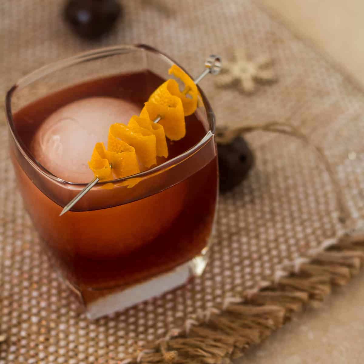 A glass of dark cocktail with a large ice sphere, garnished with a curled orange peel on a metal pick, sits on a textured brown placemat.