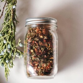 A glass jar filled with a colorful herb bruschetta mix of dried herbs and flowers sits on a light surface next to a sprig of greenery with blue berries.