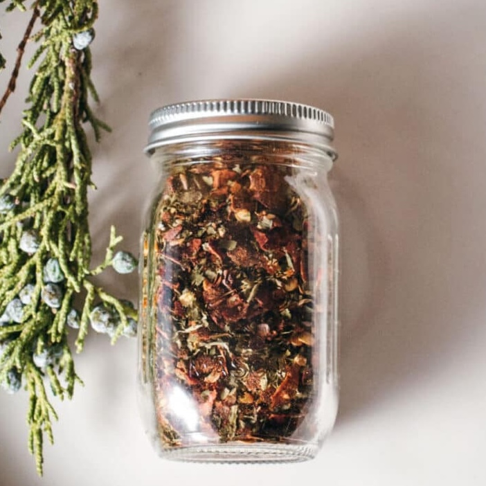 A glass jar filled with a colorful herb bruschetta mix of dried herbs and flowers sits on a light surface next to a sprig of greenery with blue berries.