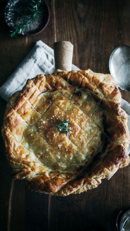 A golden, flaky pie with a crisscross pastry pattern, topped with sea salt and herbs, sits in a rustic dish on a wooden table with a napkin and small bowl nearby.