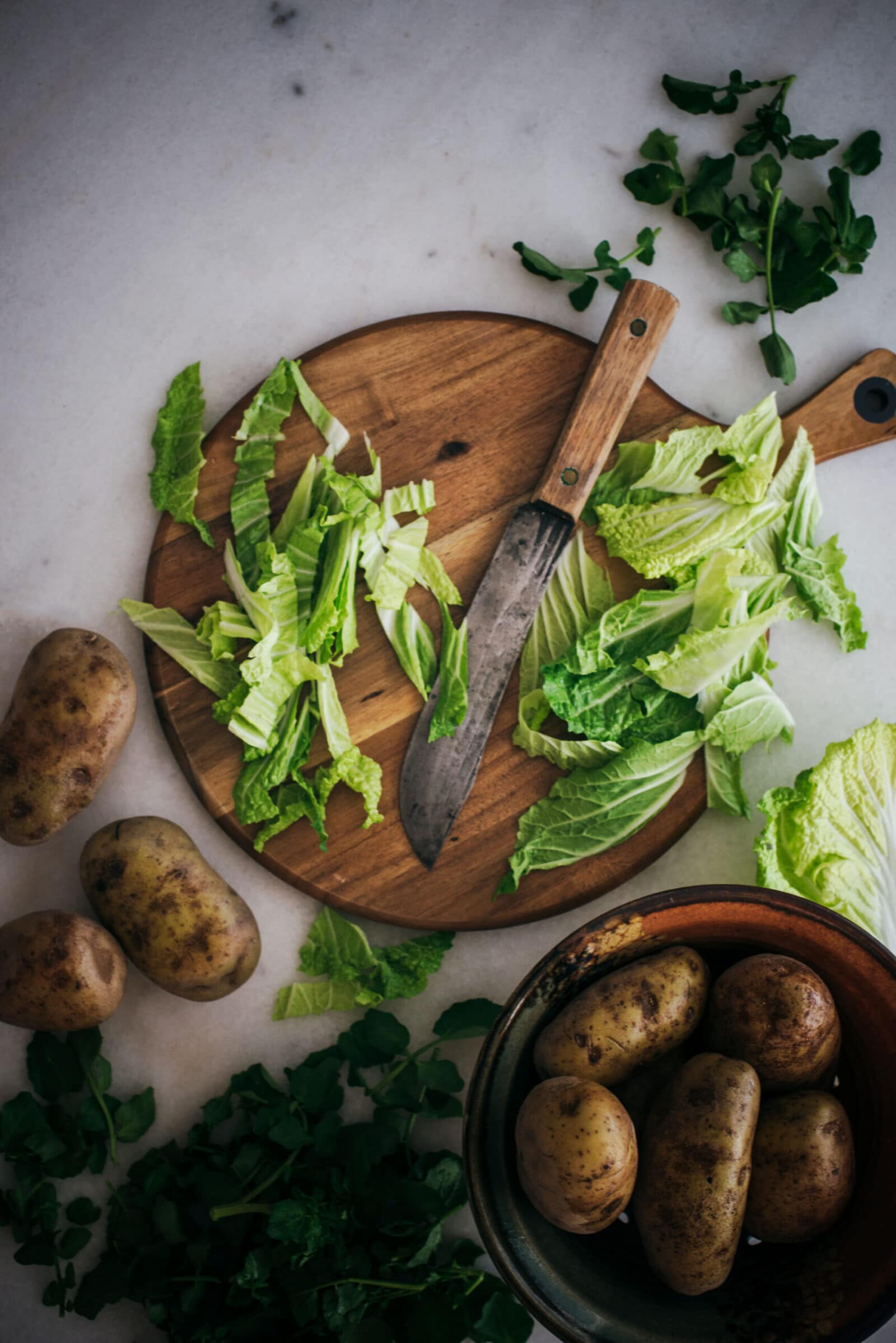 A wooden cutting board with chopped lettuce and a knife sits on a marble surface. Whole potatoes and leafy greens, classic ingredients for a Maori boil up recipe, are scattered around, with more potatoes in a bowl nearby.
