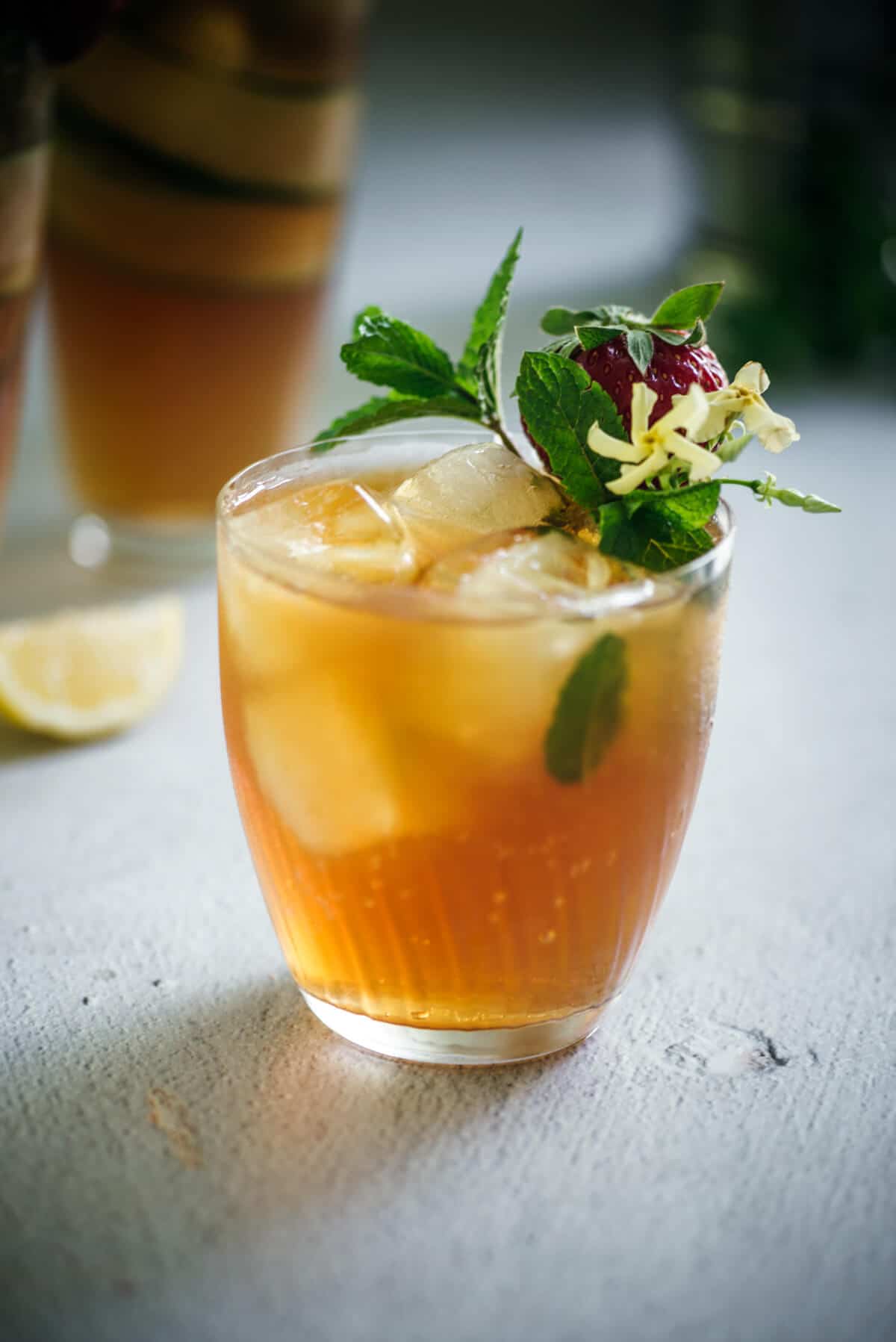 A glass of iced tea, reminiscent of a classic Pimms Cup, is garnished with fresh mint leaves, a small flower, and a berry. It sits on a light surface with a lemon wedge blurred in the background.