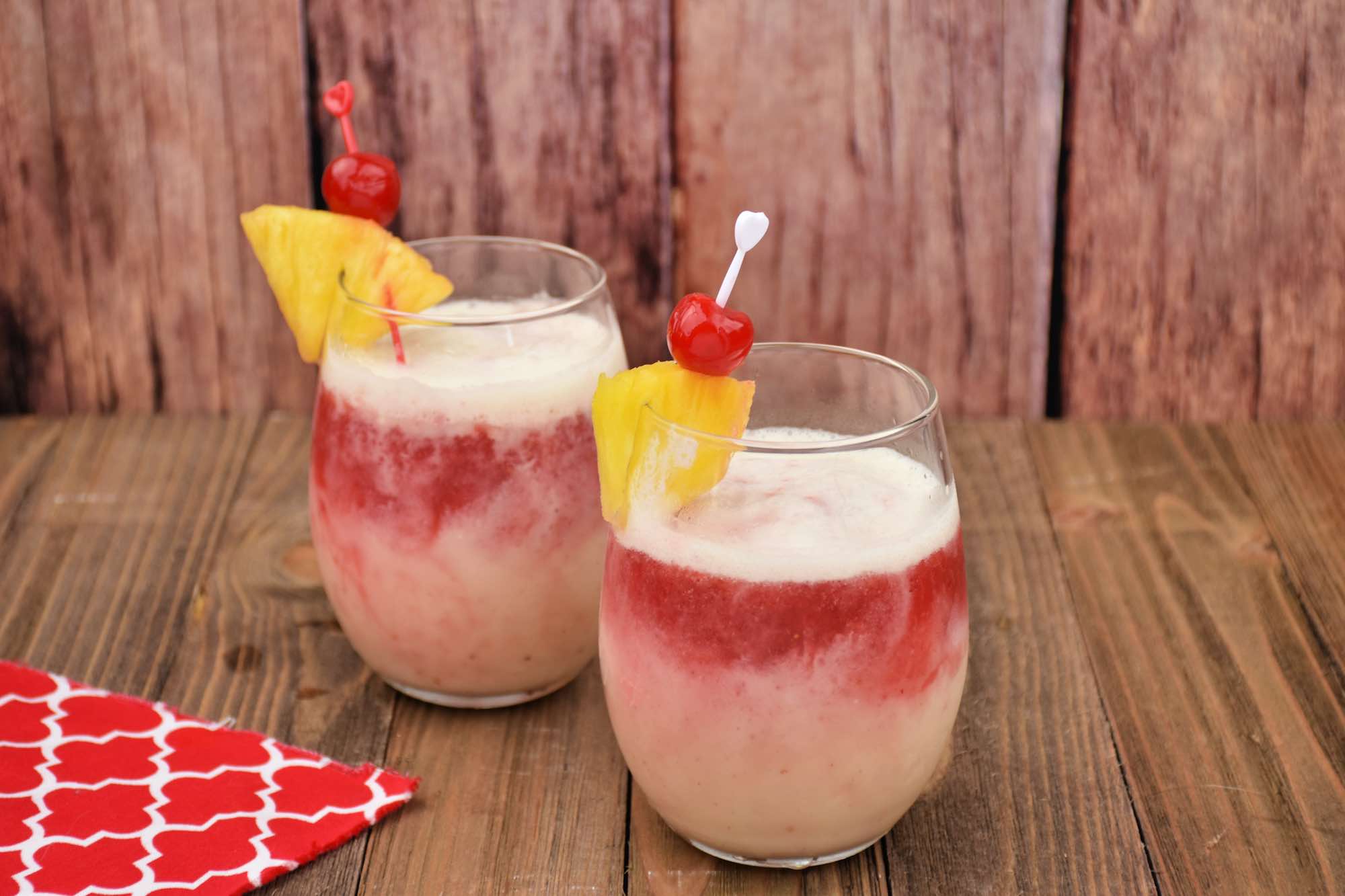 Two glasses of a layered red and white tropical drink, garnished with a pineapple slice and a cherry on a pick, set on a wooden surface with a red and white patterned napkin beside them.