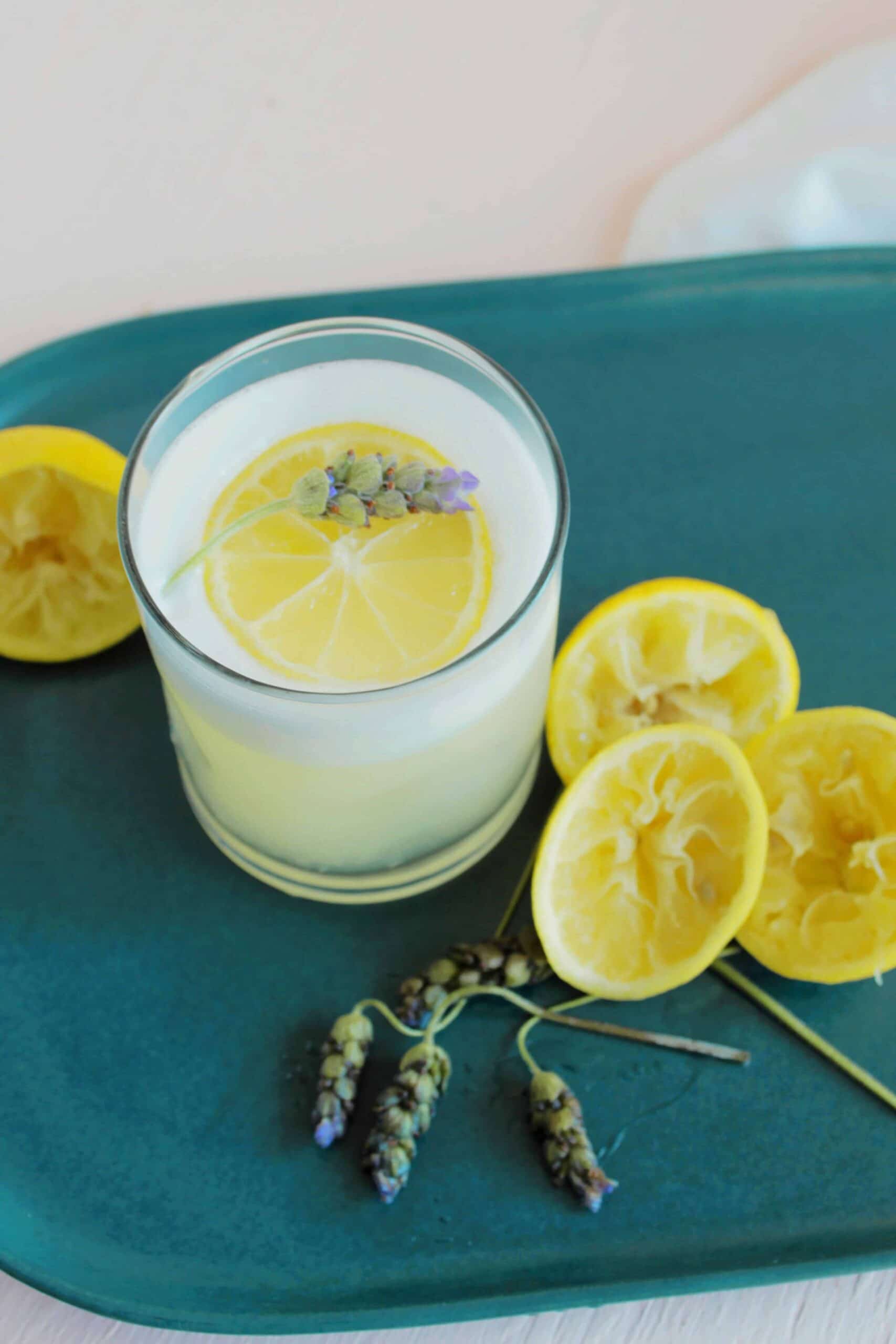 A glass of lemonade garnished with a lemon slice and a sprig of lavender sits on a teal tray, next to several squeezed lemon halves and more lavender sprigs.