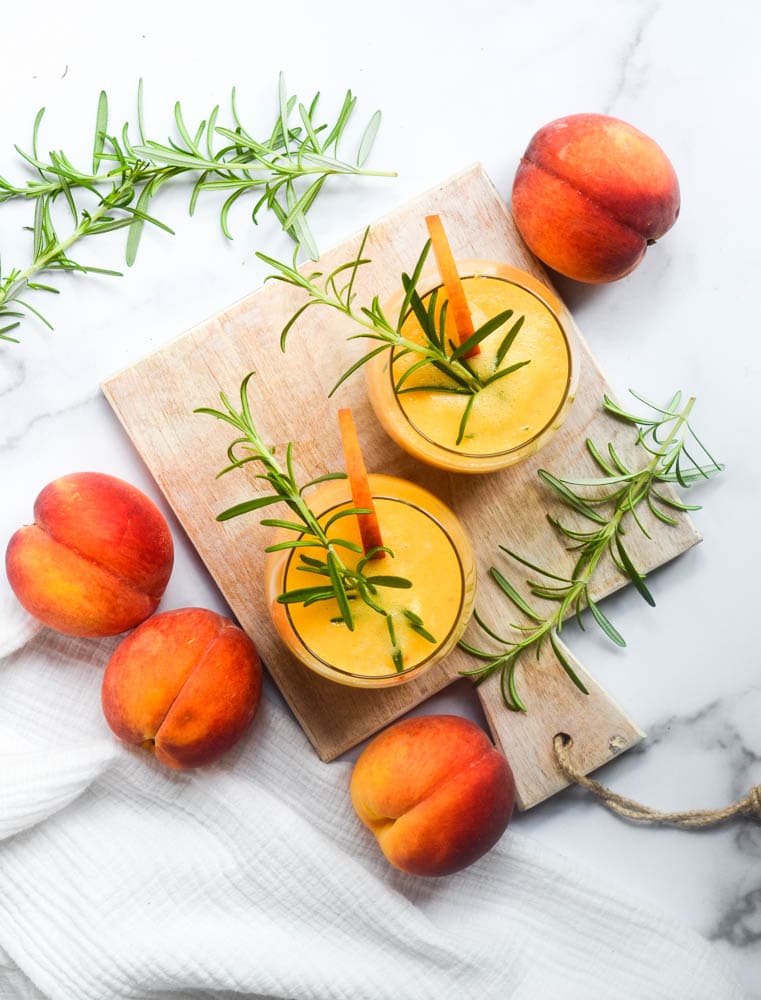 Two glasses of peach smoothie garnished with rosemary and a peach slice sit on a wooden board, surrounded by fresh peaches and rosemary sprigs on a white marble surface.