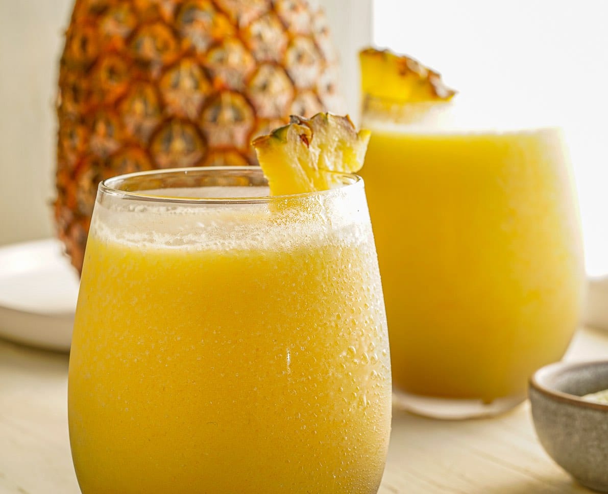 Two frosty glasses of yellow pineapple smoothie, each garnished with a fresh pineapple wedge, are set on a light surface with a whole pineapple blurred in the background.