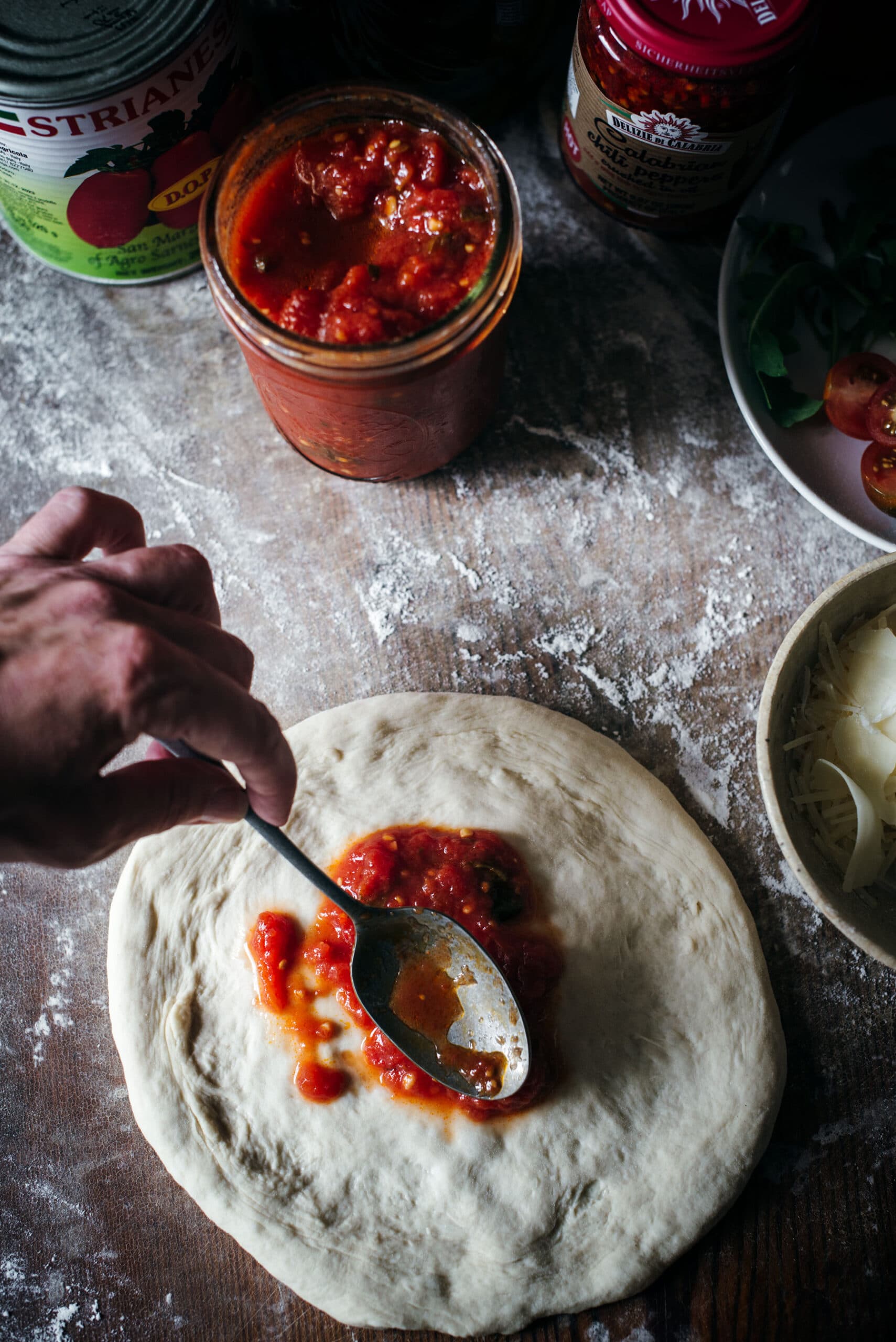 A hand spreads tomato sauce with a spoon onto pizza dough on a floured surface, surrounded by jars of sauce, cheese in a bowl, and sliced tomatoes—perfect preparation for a wood fired pizza.
