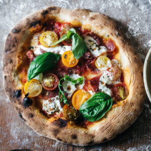 A rustic wood fired pizza topped with yellow and red cherry tomatoes, fresh basil leaves, dollops of ricotta cheese, and prosciutto on a wooden surface. The crust is golden and slightly charred.