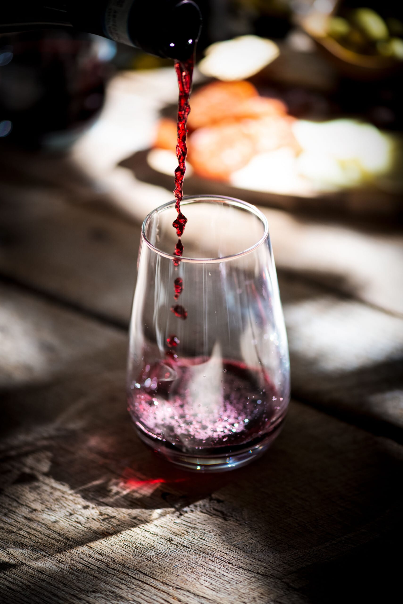 Red wine, noted for the characteristics of a good wine, is being poured from a bottle into a clear glass on a wooden table, with a blurred background featuring food items and warm lighting.