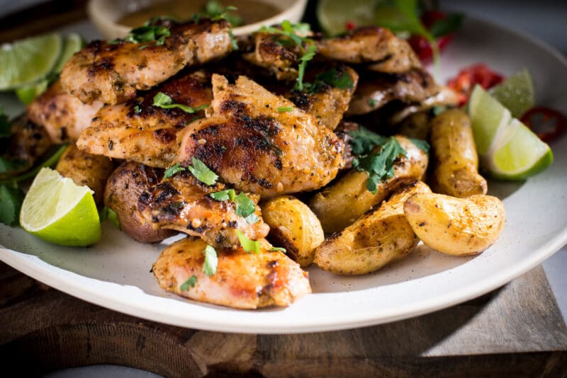 Grilled piri piri chicken pieces served with roasted baby potatoes, garnished with fresh cilantro and lime wedges on a white plate. A bowl of dipping sauce sits in the background.