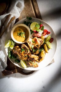 A plate of piri piri chicken, fingerling potatoes, lime wedges, red chili slices, and fresh cilantro, served with a creamy dipping sauce on a white plate atop a wooden board—perfect for fans of bold Nandos chicken flavor.