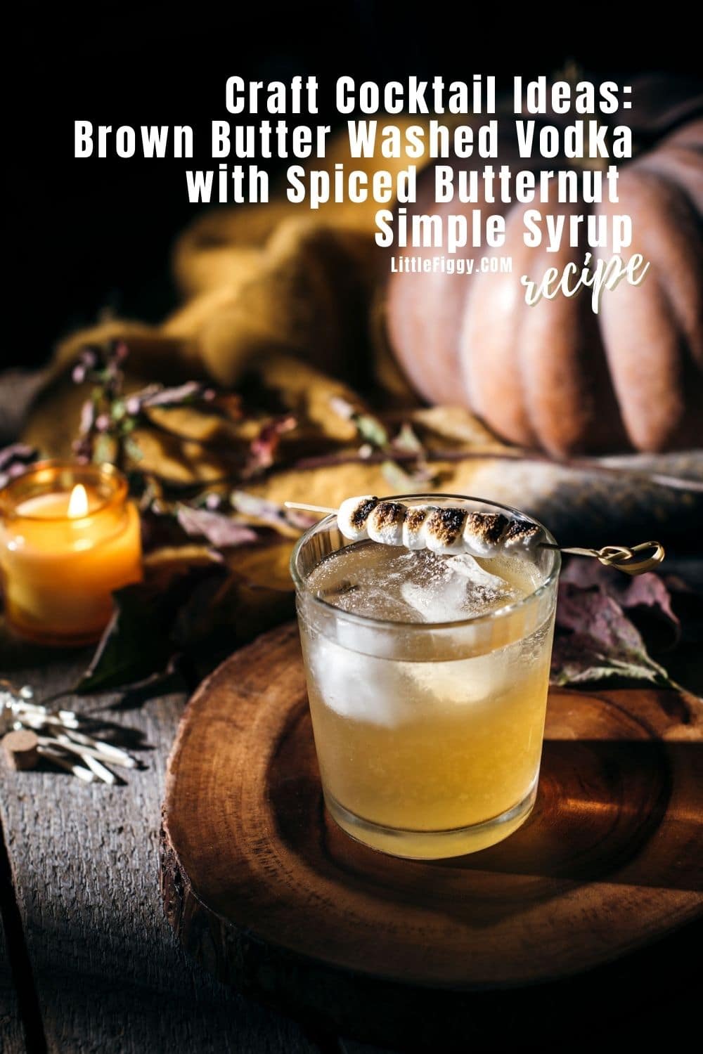 A cocktail with ice and a toasted marshmallow garnish sits on a wooden tray, surrounded by autumn leaves, a burning candle, and pumpkins—perfect for fans of easy vodka cocktails featuring brown butter washed vodka and spiced butternut syrup.