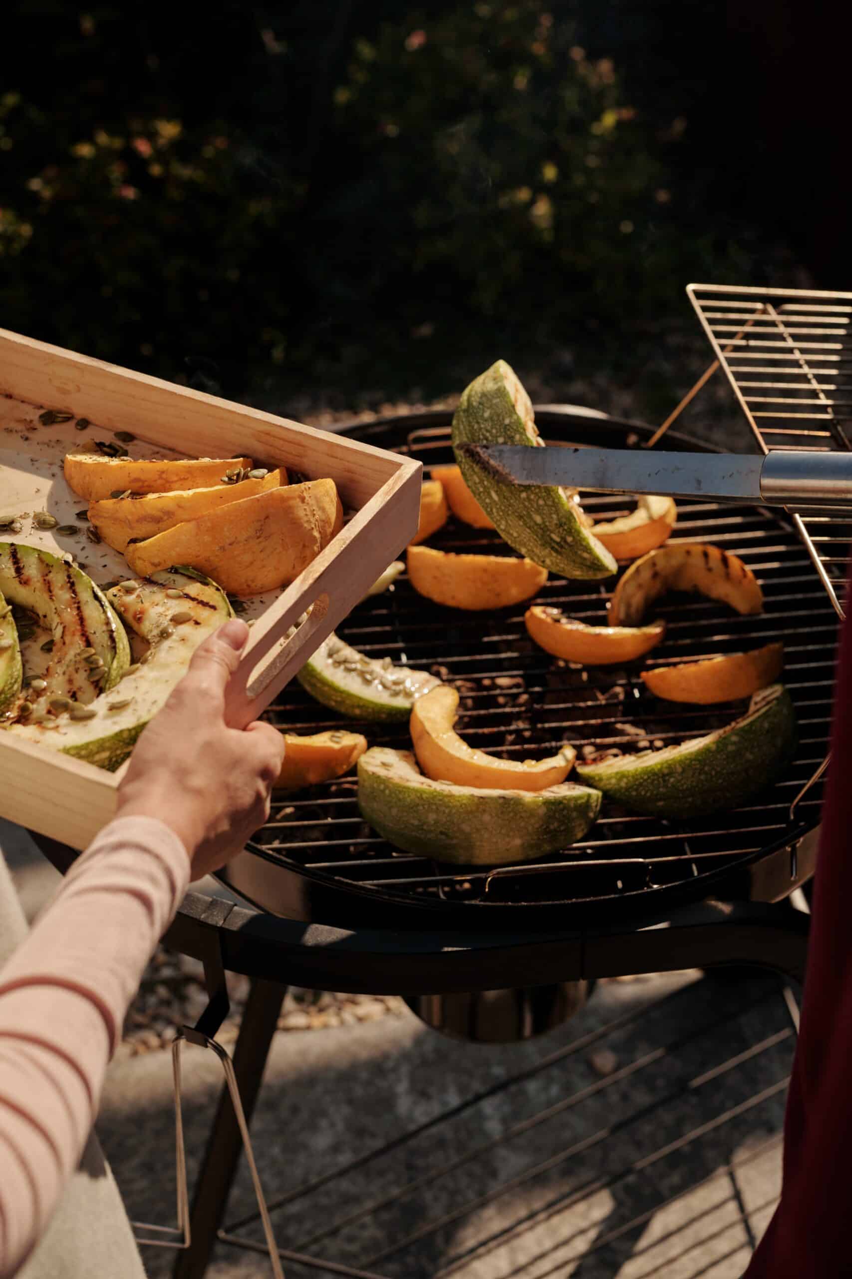A person demonstrates how to grill fruits and vegetables by placing sliced squash and zucchini onto a grill outdoors, joining more pieces already cooking on the grate.
