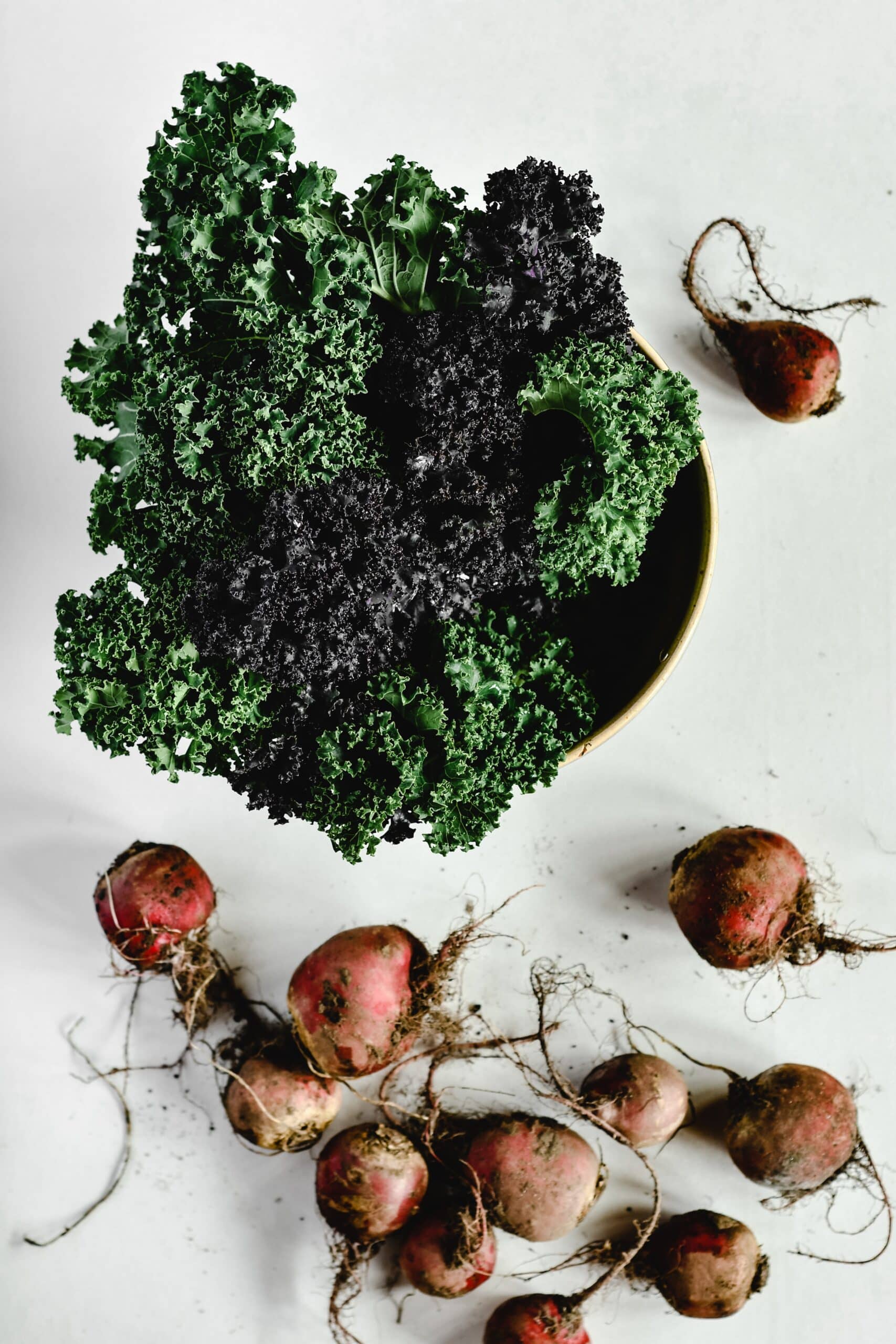 A bowl of curly kale, one of the popular superfoods, sits on a white surface next to several red beets with roots and dirt still attached.