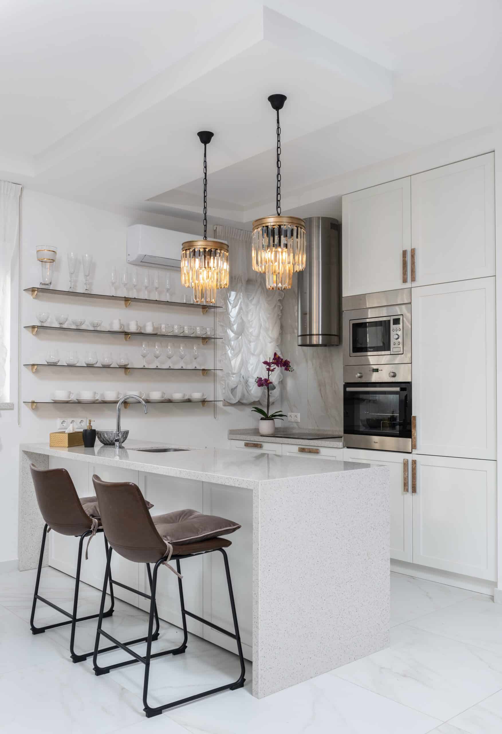 A modern white kitchen featuring a small island, two brown barstools, pendant chandeliers, open shelves with glassware, built-in ovens, and a potted orchid—a perfect showcase for stylish kitchen upgrade ideas.