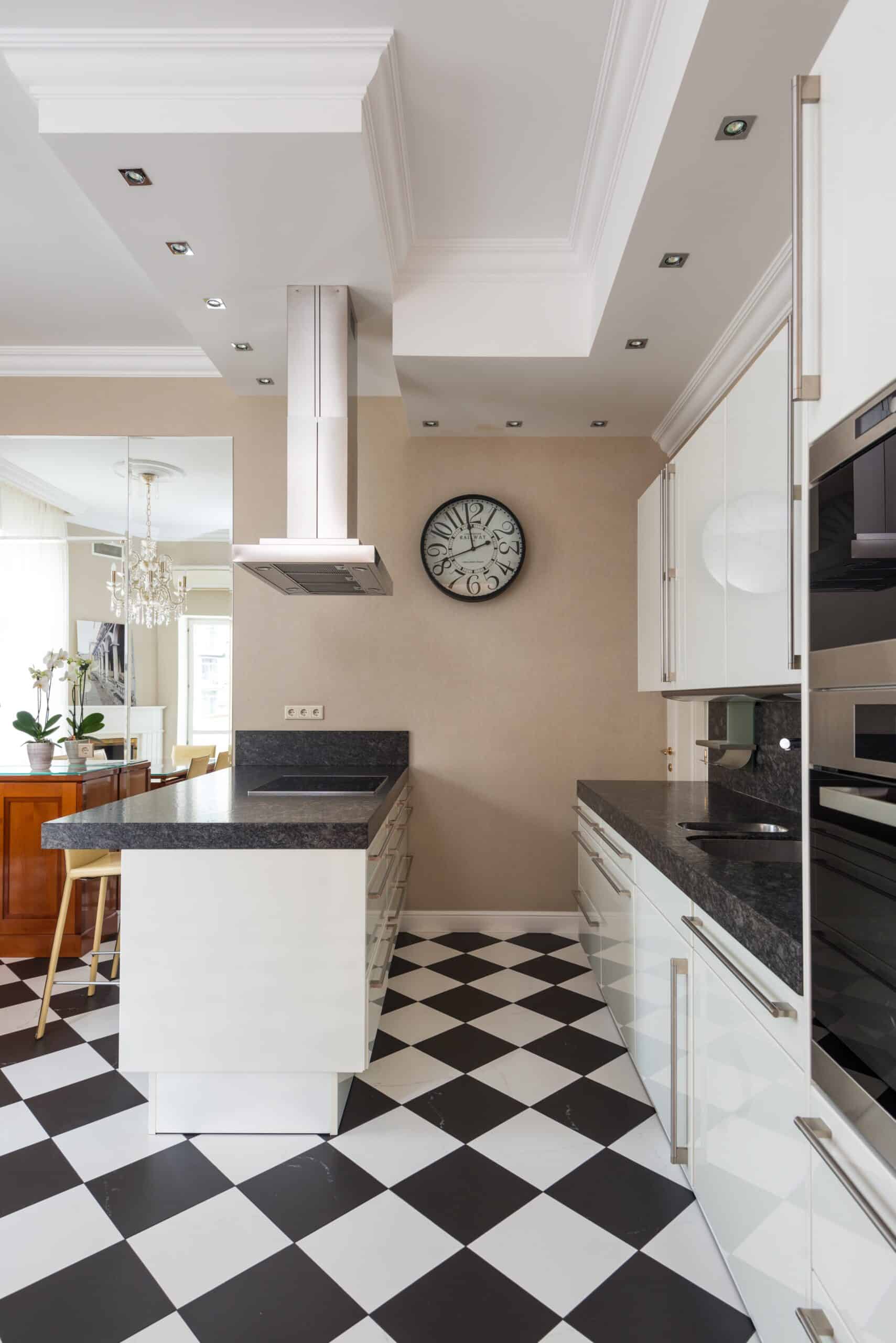 Modern kitchen with black and white checkered floor, white cabinets, dark countertops, central island with bar stools, wall clock, stainless steel range hood, and sleek wall oven. Dining area with chandelier visible in the background.