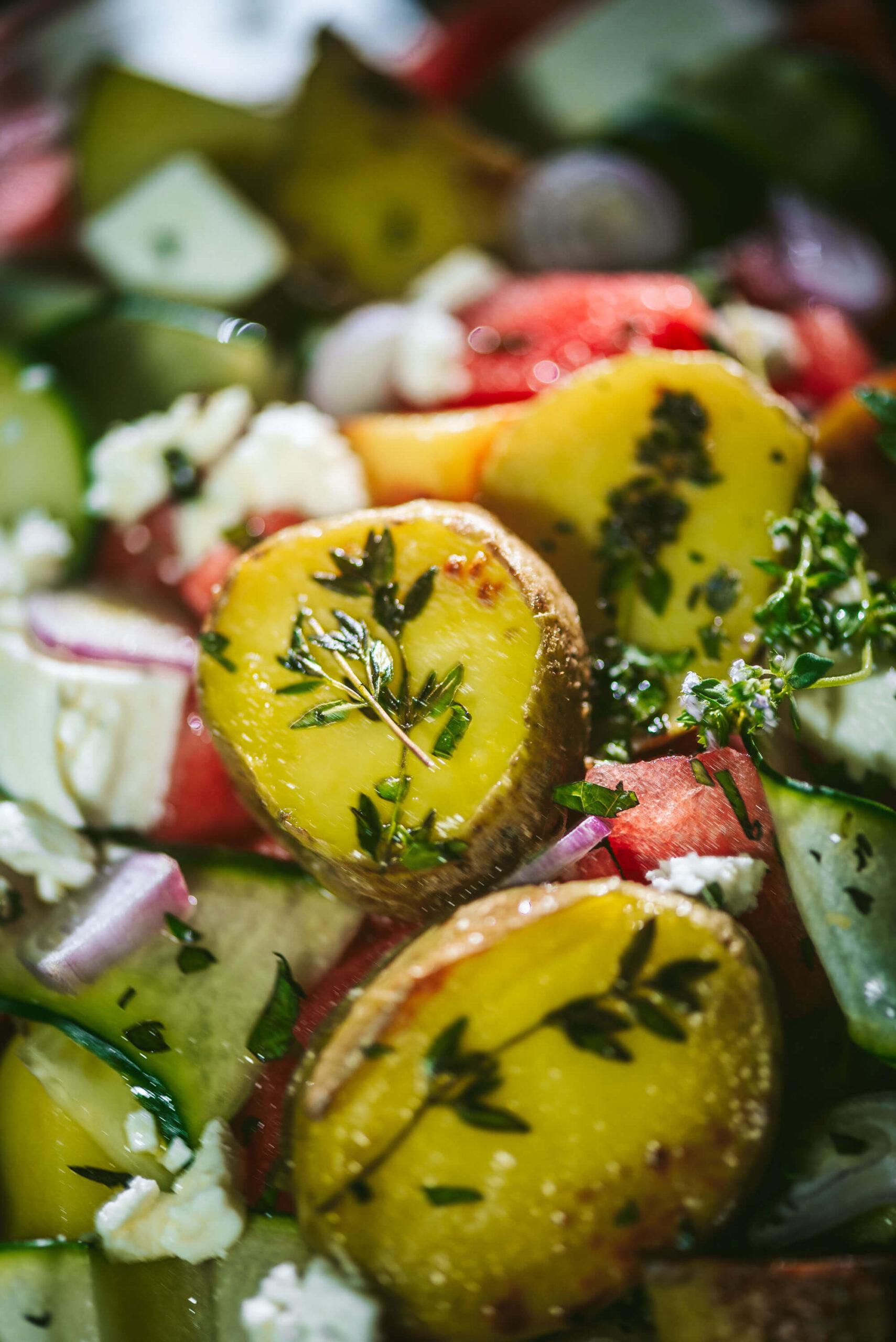Close-up of a fresh salad with roasted potatoes topped with herbs, diced cucumbers, red onions, watermelon chunks, and crumbled cheese, all in vibrant, natural lighting.