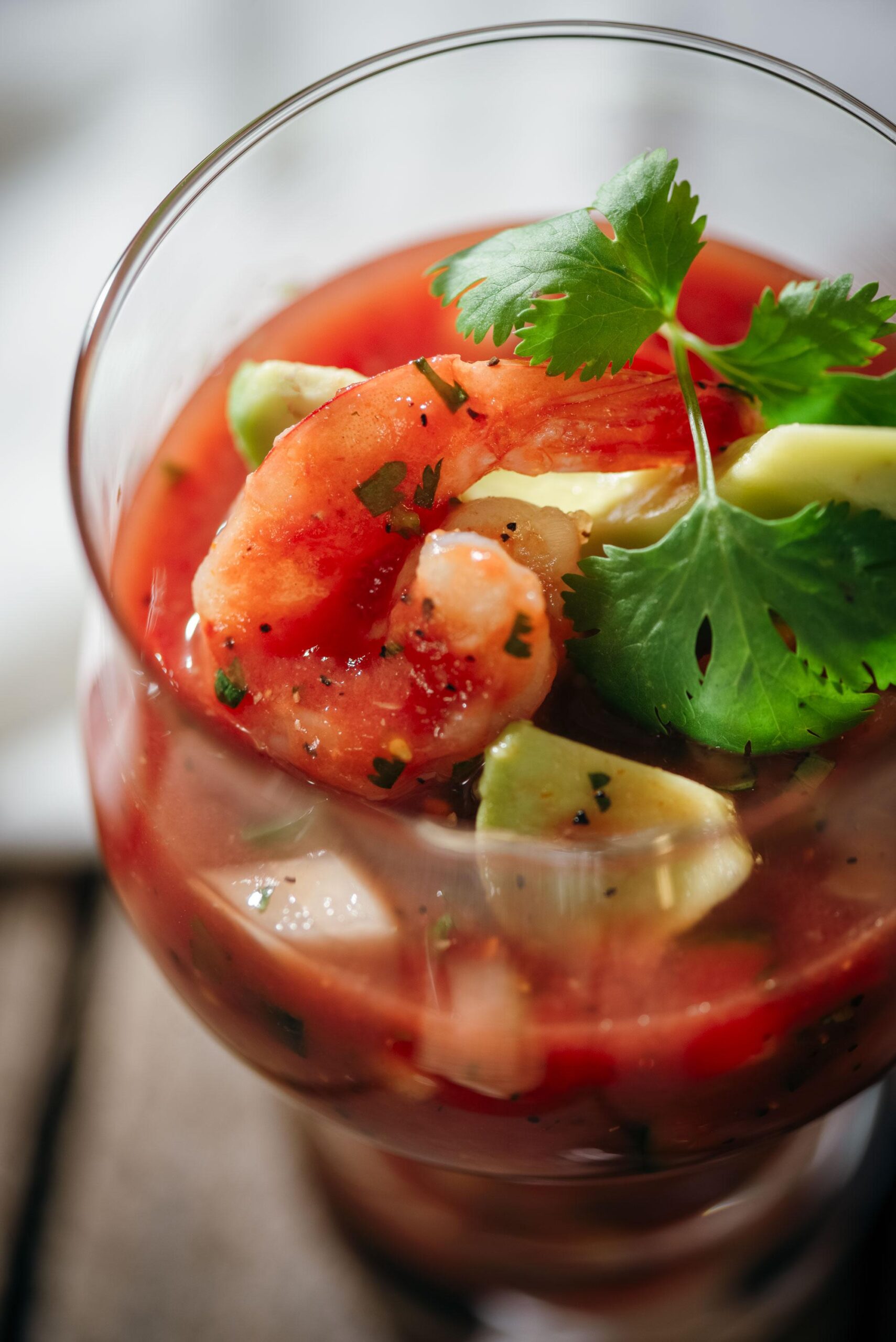 A glass filled with shrimp cocktail, garnished with fresh cilantro and avocado pieces, in a vibrant red tomato-based sauce.