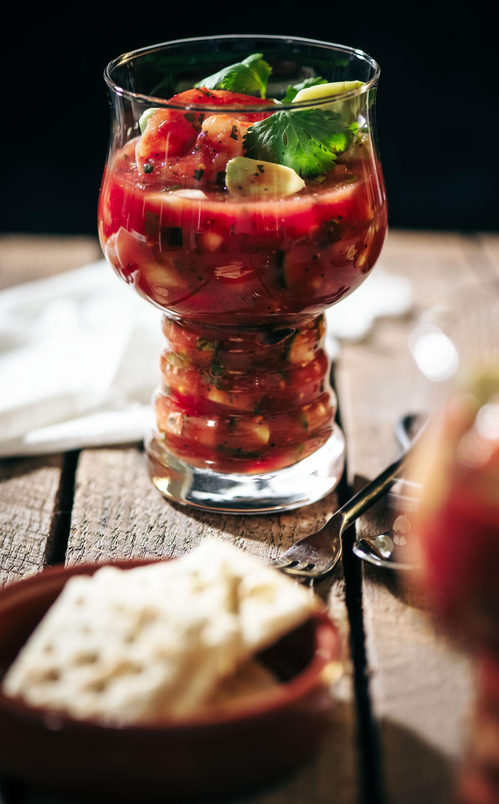 A glass filled with chunky shrimp cocktail garnished with cilantro sits on a wooden table. In the foreground, a plate with crackers is slightly out of focus. Bright natural lighting highlights the dish.