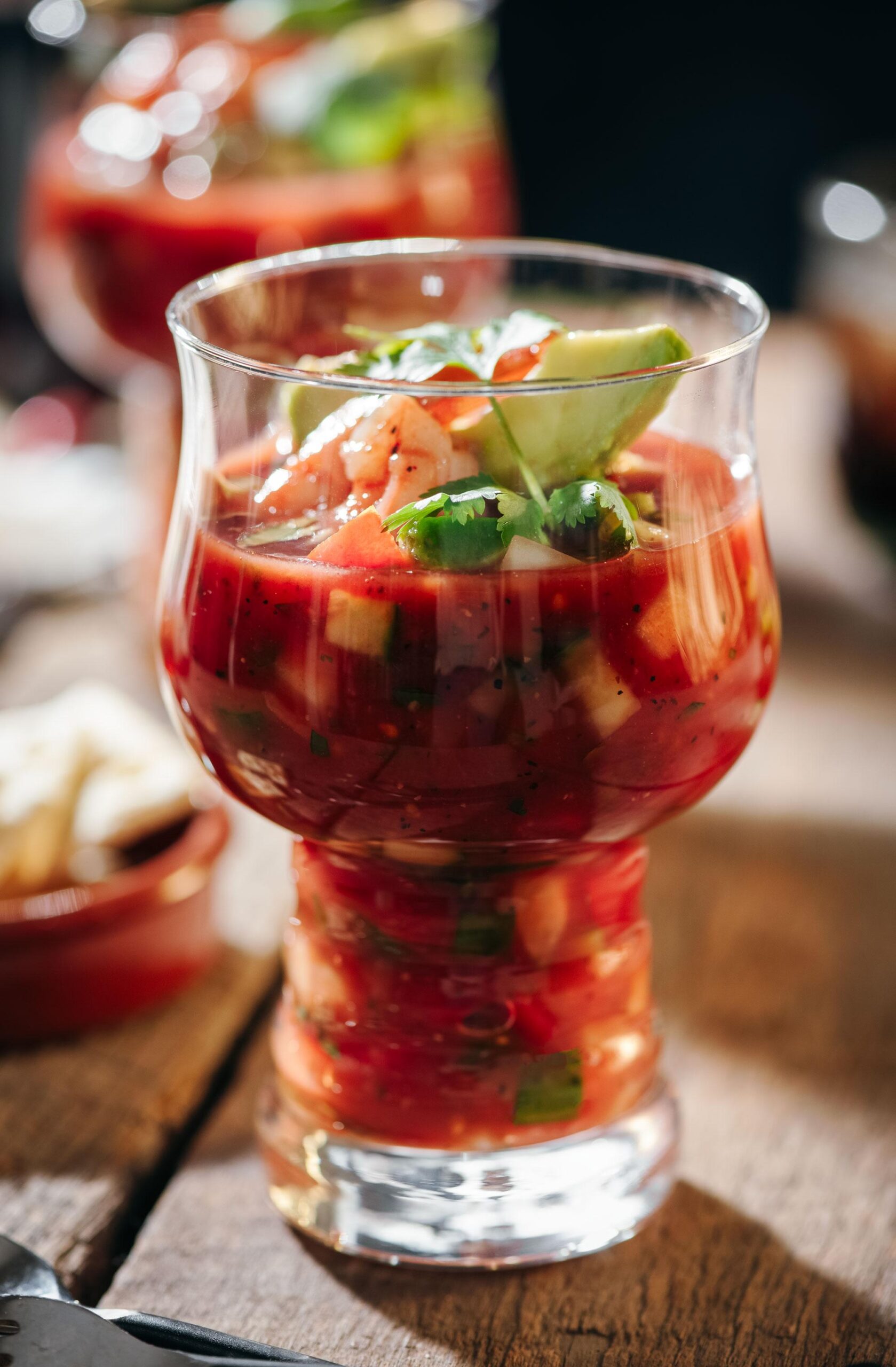 A close-up of a glass filled with shrimp cocktail in a tomato-based sauce, garnished with avocado slices and cilantro, sitting on a rustic wooden surface.