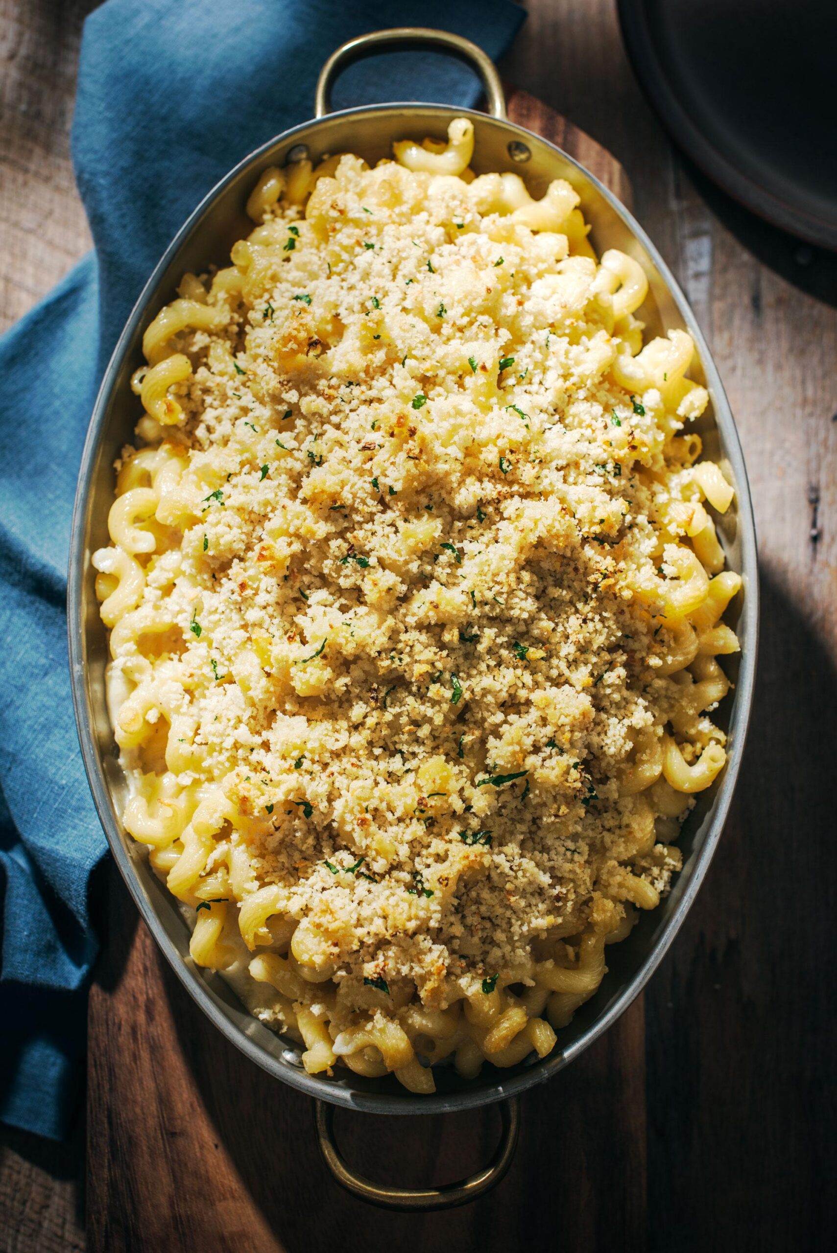 An oval baking dish filled with macaroni and cheese topped with golden, crispy breadcrumbs, placed on a wooden surface next to a blue cloth napkin.